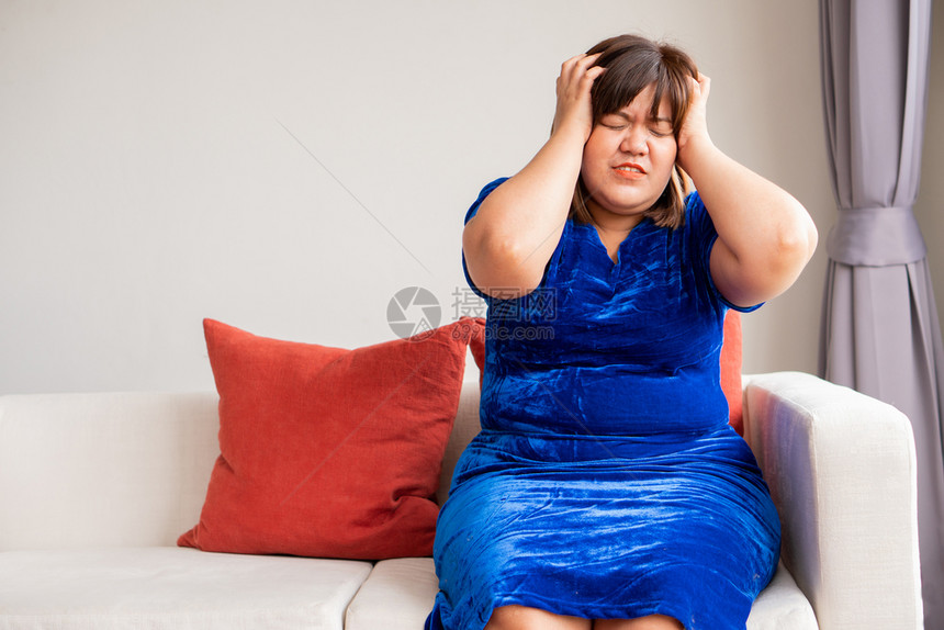 关心伤退休一位超重的亚洲妇女坐在客厅沙发上头部因偏痛而握手因为患有偏头痛疾病概念和保健护理图片