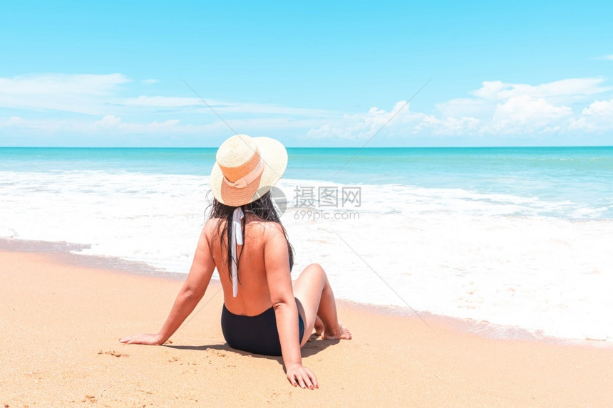 身穿泳衣和草帽的亚洲旅行者妇女背视坐在海面喷水中看洋蓝色地平线同时在节日旅行中放松美丽的吸引人泳装图片