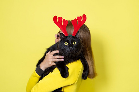 女孩逗猫猫咪穿黄色裙子的女孩拿着她心爱的毛茸滑稽黑猫戴着圣诞驯鹿角头带戴着黄色背景的圣诞猫黄色背景脸节背景
