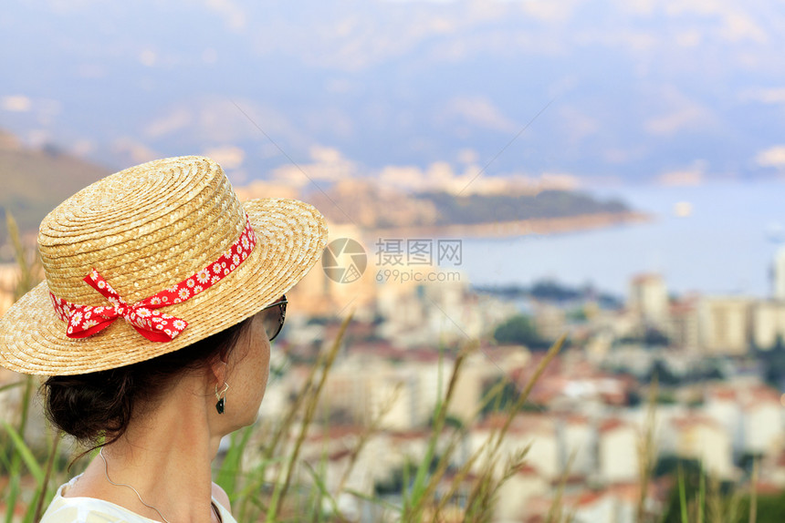 身戴草帽的年轻女子戴着莫特利红丝带和墨镜正从海湾高处和布德瓦市Budva看望着身戴草帽的年轻女子看着海港和黑山布德瓦市对比美丽的图片