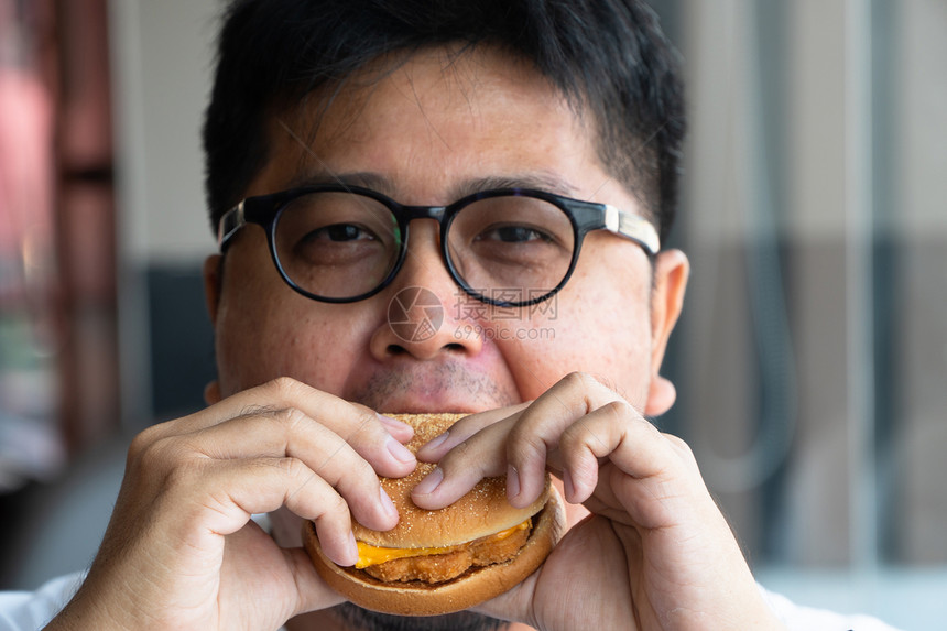 男亚洲人在快餐厅吃汉堡包享受着美味的食物穿着白色T恤和眼镜的人拿着汉堡包享受的口味笑咬饮食图片