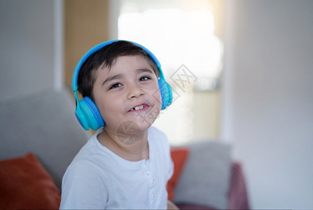 喜悦童年歌曲快乐的孩子穿着耳机仰望微笑的脸快乐男孩听音坐在沙发上玩电子游戏或在家客厅休息时的可爱基德CuteKid坐在沙发上情感高清图片素材