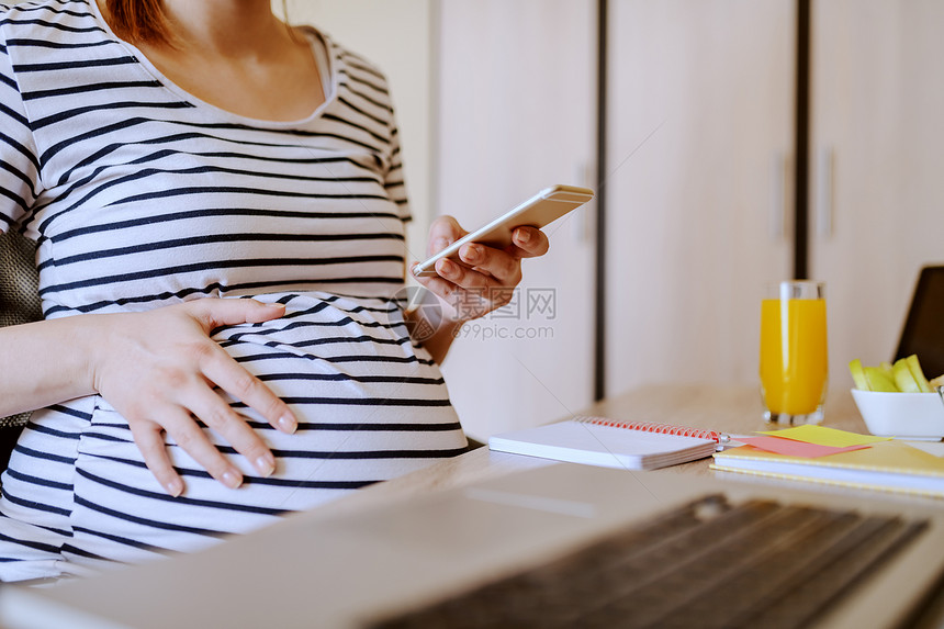 女白种年轻的孕妇在坐桌上的笔记本电脑健康早餐和带果汁的玻璃桌边时使用智能电话怀孕妇女的工作概念婴儿关心图片