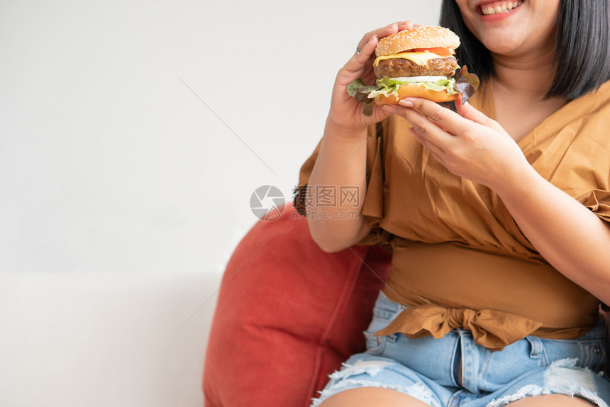 房间肉饥饿的超重妇女笑着拿汉堡包坐在客厅里她非常快乐并享受吃餐狂食饮紊乱概念面包图片