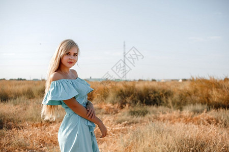 烈日炎炎发型身着浅蓝色礼服的美丽古巴年轻金发女孩站在田地上带着阳光遮的草地时髦肖像背景