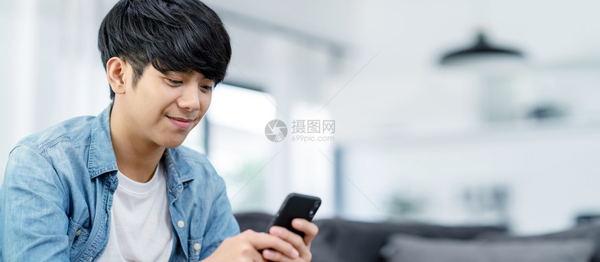 活的千禧一代技术使用智能电话在家沙发起居室微笑的亚洲男子持有并使用手机在互联网上搜索数据和社交中世纪的数据或上网搜索社会中世纪时图片