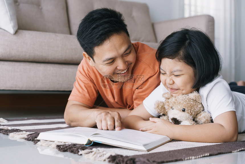 年轻的房间人们亚洲父亲和女儿正在读书同时躺她家客厅的地毯上可爱的小女孩拥抱着娃与他父亲笑得开心快乐地微笑图片