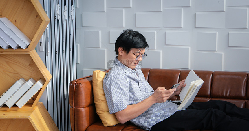 快乐的亚洲老人在家中客厅沙发上读书和使用智能手机的亚洲老年人长者肖像正在放松和幸福地阅读一本杂志校对Portnoy女士亚洲人图片