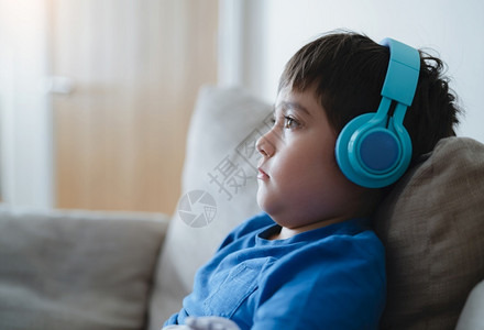 身穿耳机深思熟虑的男孩穿着蓝色T恤听音乐的学校男孩CuteKid坐在家中客厅的沙发上休息时唱歌在线的乐趣年轻的高清图片素材