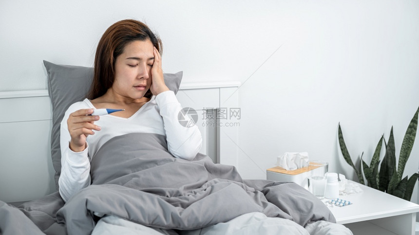 新冠亚裔妇女在用手摸脸时感到生病和头痛坐在床上的毯子同时在服药前检查温度计以测量体使用手碰脸时感到恶心和头痛随意的封锁图片