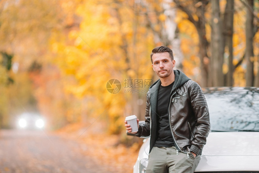 在秋天喝咖啡的快乐男子乘坐汽车旅行青年男子在秋天公园户外用手机喝咖啡的青年男子一次十月外部图片