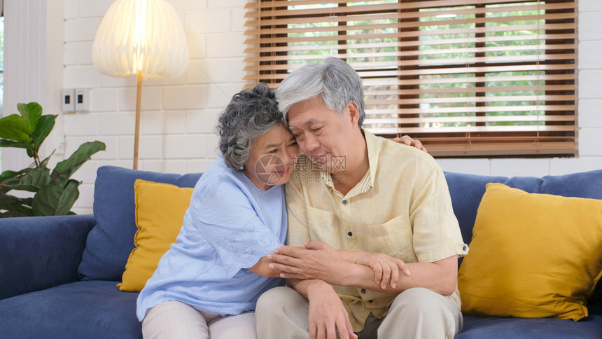 担心的关怀年长亚裔夫妇在家中客厅坐沙发上时因情绪低落而互相安慰享受老年退休生活方式辅助图片