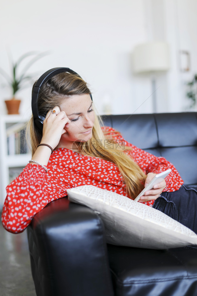 活的吸引人内部听音乐时戴耳机的快妇女使用智能手机坐在室内住宅起居沙发上的听音乐图片