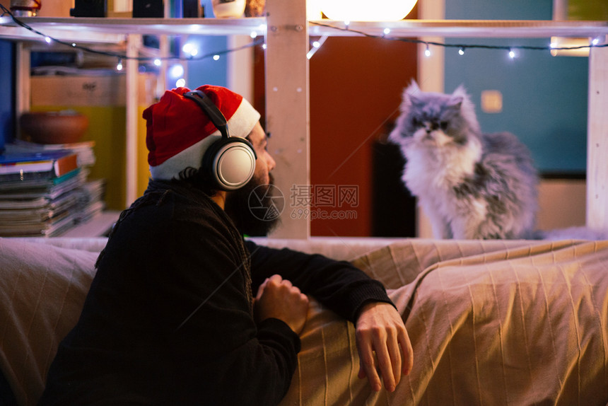一种带着耳机和圣诞帽的年轻人看着他的猫在沙发上有装饰灯光红色的礼物图片