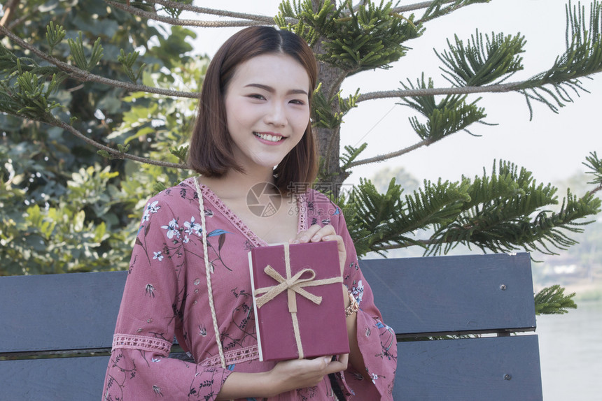 红色的年轻青女快乐微笑在公园手里拿着礼物盒坐周年纪念日图片