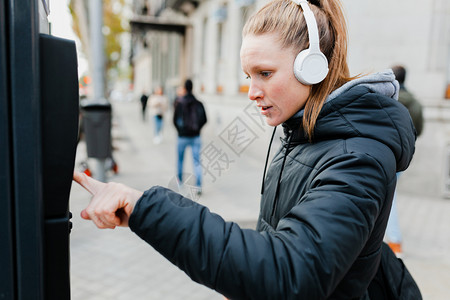 年轻的严肃快乐年轻金发美女在城里付停车费脸上戴着耳机的夹克脸型惊讶的图片