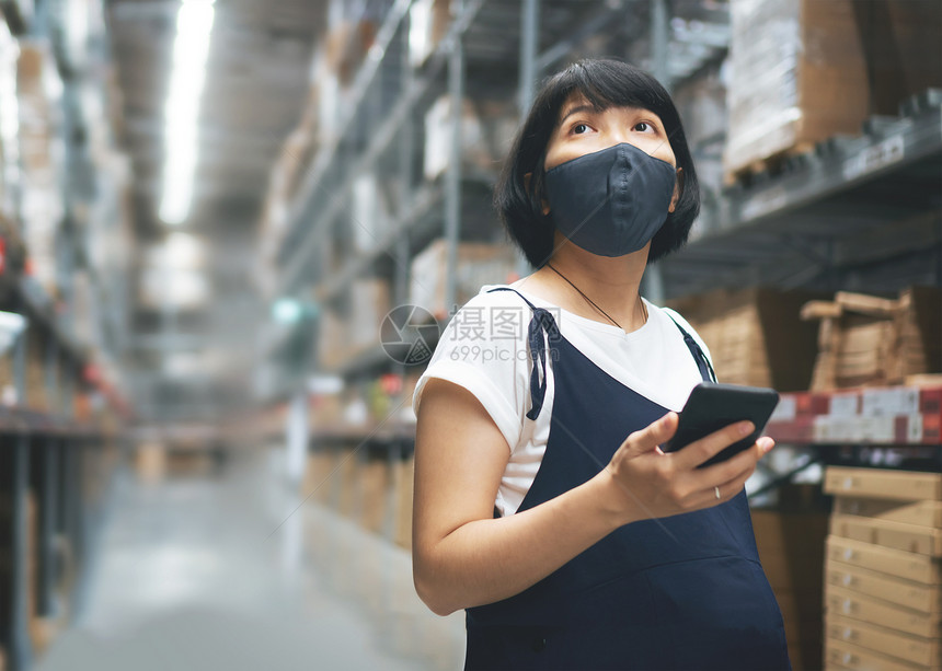 创业者身戴保护面罩的女企业主在仓库商店持有智能手机而怀孕的年轻亚洲妇买家则使用电话和看商场的货架在仓库店里装有智能手机店铺购物面图片