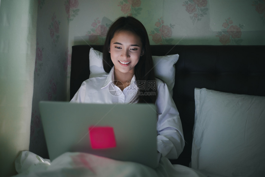 男人互联网亚裔妇女用笔记本在床上夜间工作她微笑并享受在家工作移动的图片