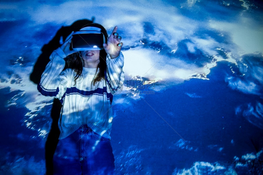 肖像妇女使用虚拟现实头盔在带有多彩投影器光照明渗透概念的交互式技术展览中手动移女使用虚拟现实头盔小工具虚拟的图片