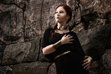 神话美丽的化妆品女孩身着黑色外衣和古董珠宝背面有岩石墙女神年轻的图片