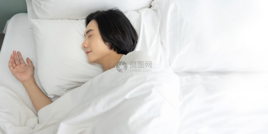 年轻人早上睡在白床做梦快乐笑的亚裔男享受躺在舒适软床铺上在家眼闭着的复制空间中躺在家里的卧室安详放松平静的图片