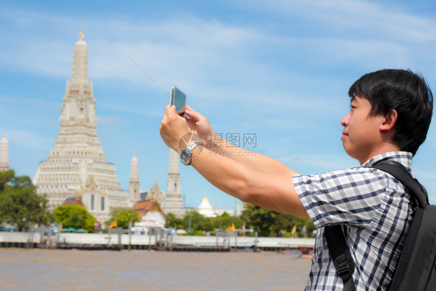快乐的旅游男人在泰国河上站着自拍旅行者拿着手机拍摄自己与泰国的WatArun一起微笑的照片男是背包黑色用智能手机拍照宝塔图片