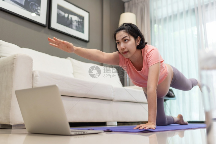 福利身体在线的年轻母亲通过笔记本电脑在沙发附近的亚洲孕妇身穿粉红色衬衣在家里伸展着粉色衬衫用在线膝上笔记本电脑进行瑜伽图片