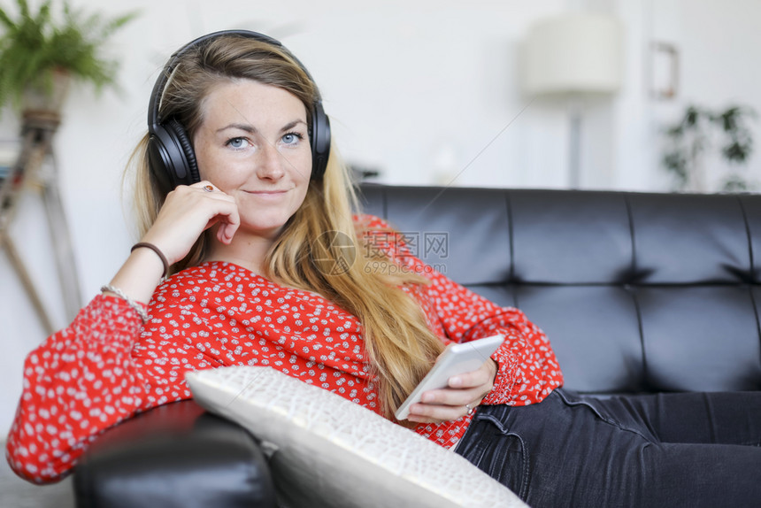 互联网听音乐时戴耳机的快女人坐在室内客厅沙发上的摄影机拿着耳看镜头玩房间图片