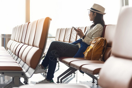 坐着亚洲人快乐的女旅行者佩戴眼镜和帽子在机场等待度假航班时却带着智能手机享受一顶聪明的帽子寻找无线科技假期无线上网高清图片素材