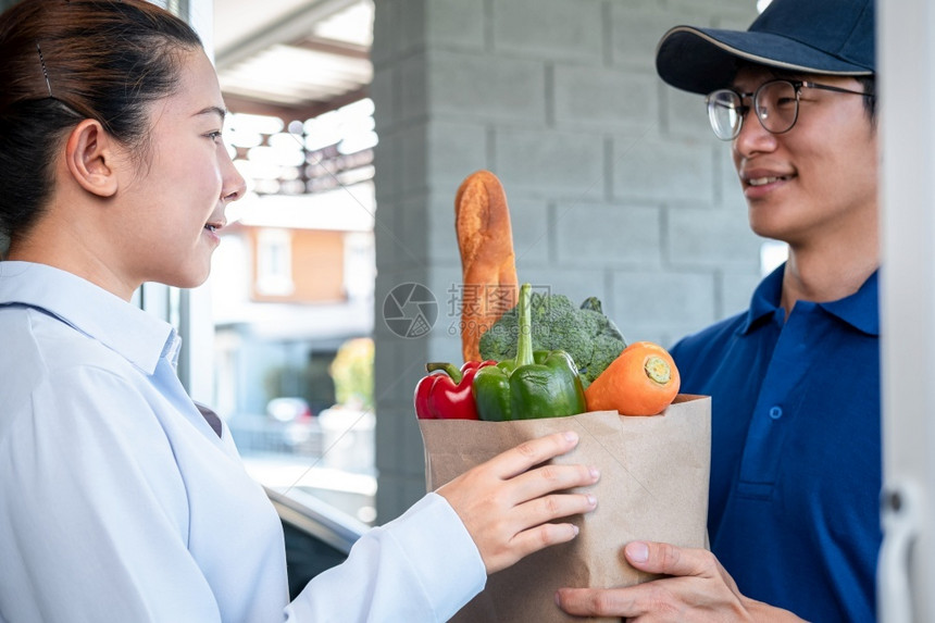 身穿蓝色制服的亚洲送货男子在Corona共19流行期间服务的房子前面向女顾客提供食品水果和蔬菜杂货袋保持邮递员快速地图片