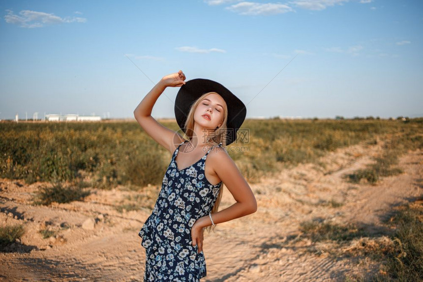 白种人村庄一位年轻美丽的caucasian金发女孩的肖像她穿着蓝色裙子印花粉和黑帽子站在沙乡村公路上夏天日落时靠近葡萄田奢华图片