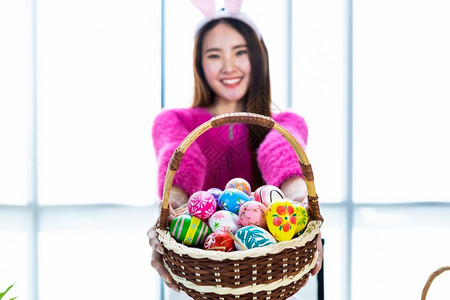 女孩兔子美丽的幸福复活节概念穿着兔子耳朵的亚洲快乐女青年士戴着兔子耳朵的抽象模糊与焦点节目集中展示拿着一篮子和多彩的复活节鸡蛋在白衣室背背景
