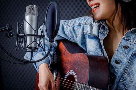 一种生产快乐的笑容年轻亚洲女发声员戴着耳机吉他在专业制片室录麦克风前的歌声笑玩愉快高清图片素材