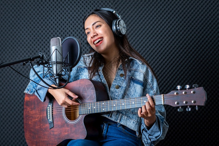 吉他手声乐专业的一位年轻亚洲女歌手戴着耳机在专业工作室录制麦克风前的歌声她笑得很高兴他开心一个年轻的亚洲女发声员戴上耳机背景图片