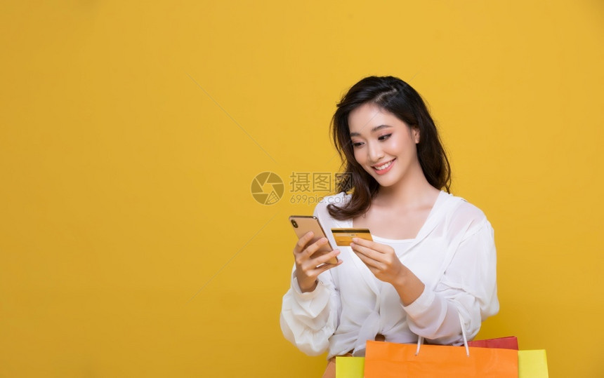 亚洲人买方美丽的年轻女微笑着快乐的容她持有信用卡使智能手机在网上購物与购袋一起买黄色背景幸福消费主义销售和人购物概念un漂亮的图片