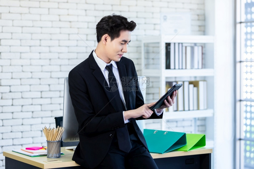现代的一位愉快乐心情一个成功的亚洲青年商人在办公室用智能手机和笔记本电脑做木制桌底背景工作一种沟通图片