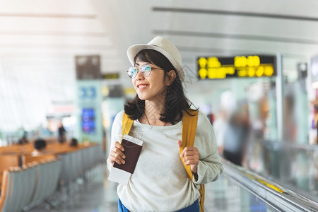 人们穿微笑快乐的亚洲妇女戴眼镜黄色背包帽的子持有飞行机票和护照同时等待机场大厅的飞起笑一国际的高清图片素材