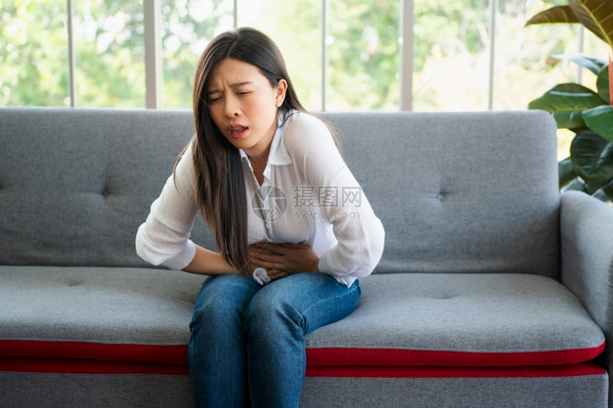 恶心亚洲人下午坐在沙发上并忍受腹部因月经泻或消食不良而痛的腹部疼不快亚裔妇女坐落在沙发上忍受着腹部疼痛疾病和保健概念图片