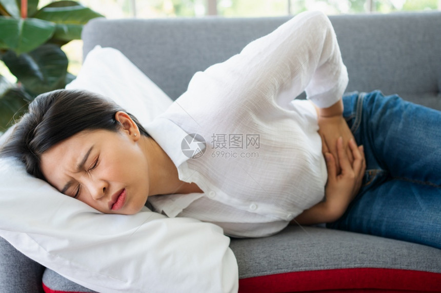 坐在沙发上并忍受腹部因月经泻或消食不良而痛的腹部疼不快亚裔妇女坐落在沙发上忍受着腹部疼痛疾病和保健概念恶心坏的时期图片