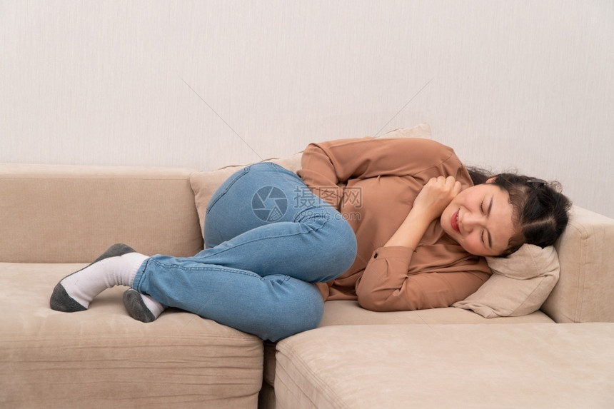 抽筋坐着在沙发上并忍受腹部因月经泻或消食不良而痛的腹部疼不快亚裔妇女坐落在沙发上忍受着腹部疼痛疾病和保健概念伤害图片
