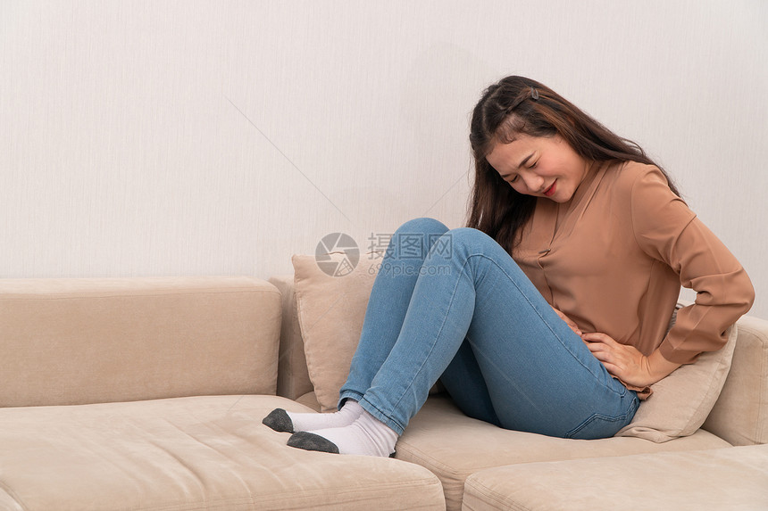 坐在沙发上并忍受腹部因月经泻或消食不良而痛的腹部疼不快亚裔妇女坐落在沙发上忍受着腹部疼痛疾病和保健概念抽筋或者来了图片
