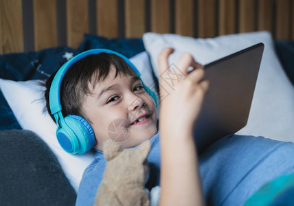 年轻的快乐坐在床上无线耳机监听音乐在平板电脑上玩游戏穿着蓝睡衣微笑的孩子早上在睡觉室放松然后去学校上笑着生娱乐高清图片素材