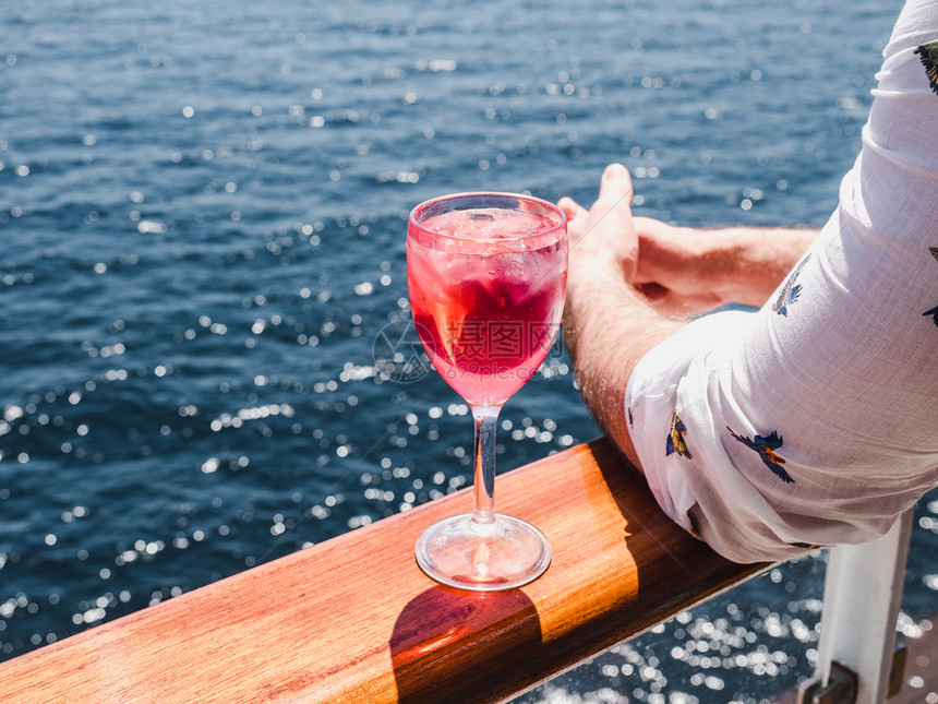 冰庆典红色的在蓝海浪背景下游轮露天甲板上盛着一杯美丽的粉红葡萄酒时装男子侧观特近休闲和旅行概念暇和旅游概念持有美丽粉红葡萄酒的人图片