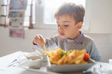 小男孩吃薯条手小可爱的caucasiansian小男孩的肖像他吃薯条片在餐厅的桌上或在家中三四岁坐在桌边白天子旁边可口儿背景