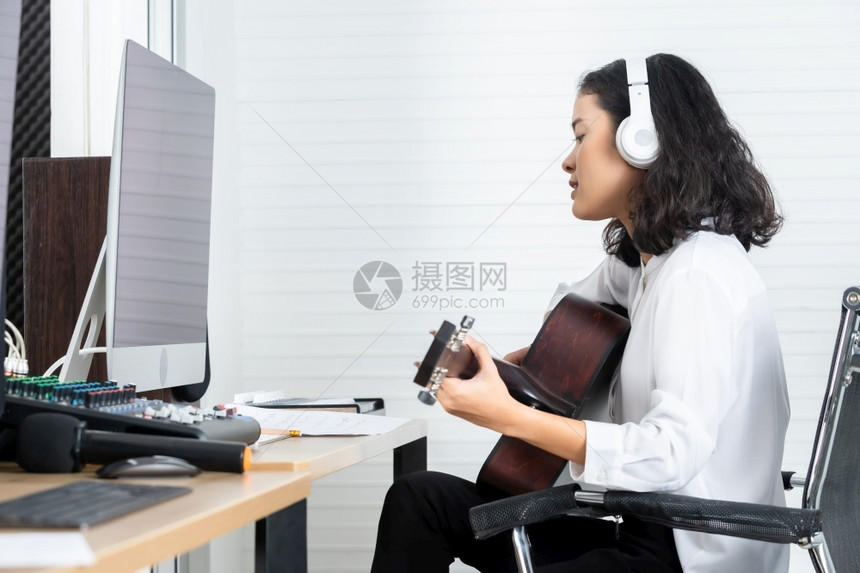 专业音乐家亚洲女青年发声员佩戴耳机弹吉他在专业演播室录制和编辑声音的软件计算机监视器的一首歌录音带记岩石电子的吉他手图片