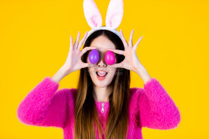 微笑复制象征活节的概念快乐笑亚洲年轻女穿着兔子耳朵的年轻女士在她眼前举着一个多彩的复活节鸡蛋在黄色空版间工作室背景上被孤立图片