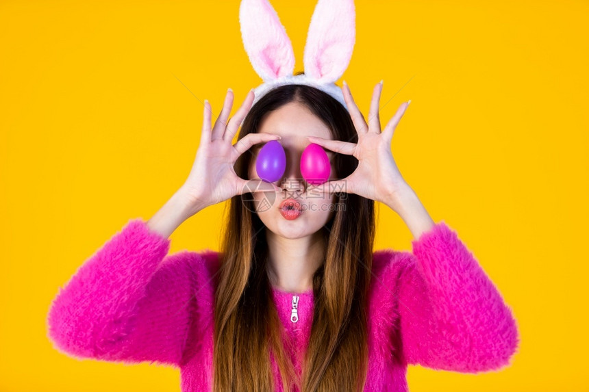 串烧黄色的复活节概念快乐笑亚洲年轻女穿着兔子耳朵的年轻女士在她眼前举着一个多彩的复活节鸡蛋在黄色空版间工作室背景上被孤立宗教图片