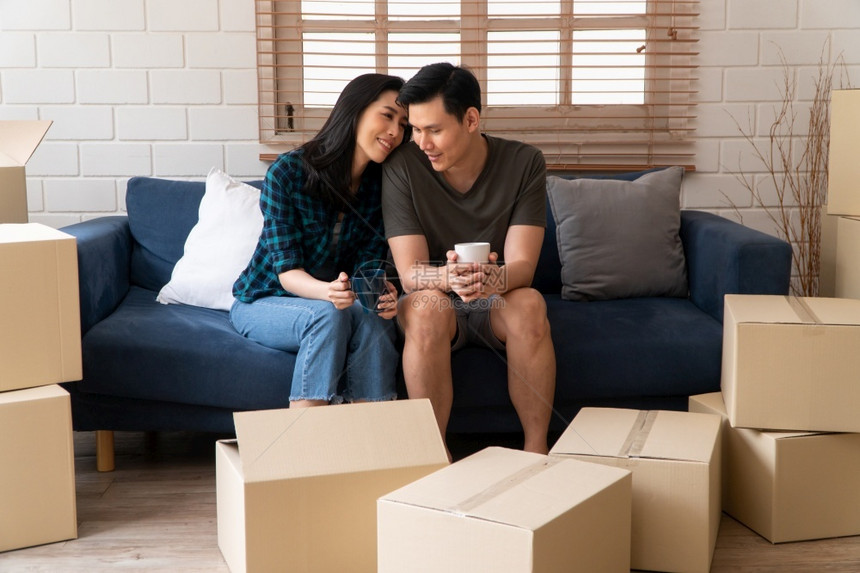 纸盒幸福的亚裔年轻夫妇拿着咖啡杯坐在沙发上休息新婚夫妇开始生活的第一天搬到新房子后构想在新婚夫妇开始生活房主家图片