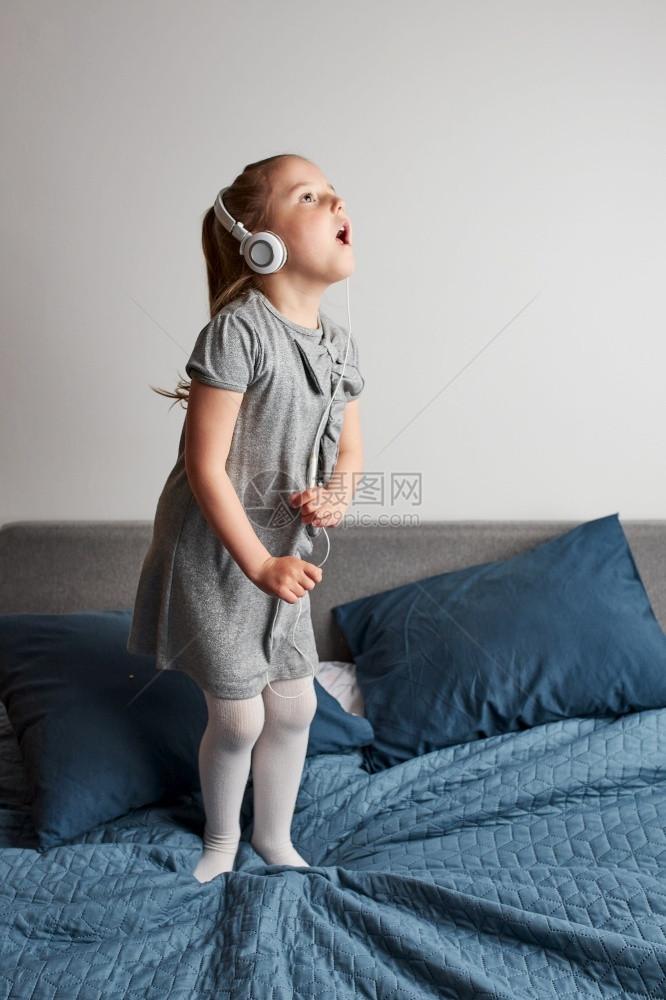 室内的乐趣美丽小女孩唱着带耳机的歌唱模仿自己一个真正的歌手Child跳舞玩乐在家里卧室的床上听音乐图片