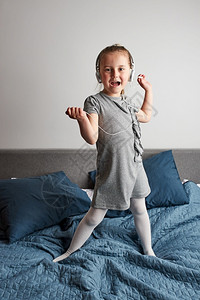 保持孩子乐趣小女唱着带耳机的歌唱模仿自己一个真正的歌手Child跳舞玩乐在家里卧室的床上听音乐模拟高清图片素材